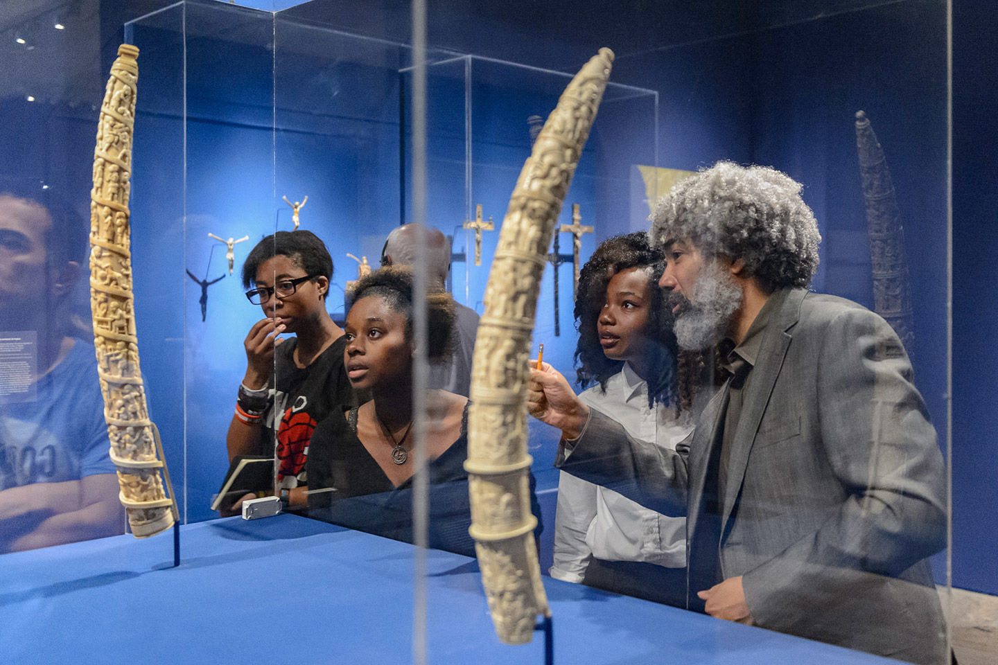An instructor and group of teenagers look with intent and awe at carved elephant tusks in glass cases within a spot-lit and blue-walled gallery